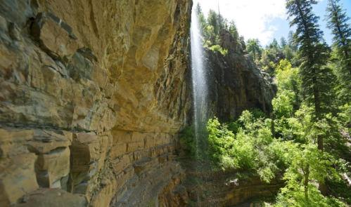 A waterfall cascades down rocky cliffs surrounded by lush green trees and bright blue skies.
