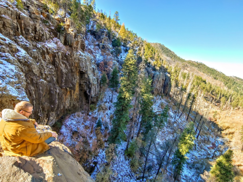 A person sits on a rocky ledge overlooking a snowy canyon surrounded by tall trees and mountains under a clear blue sky.