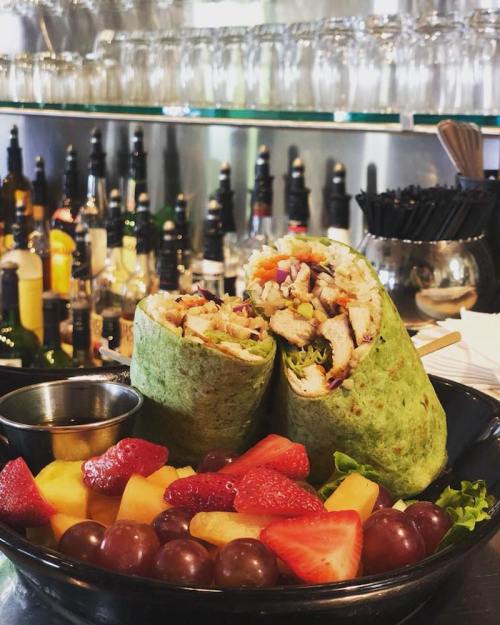 A green wrap filled with chicken and vegetables, served with a side of mixed fruit in a bowl, against a bar backdrop.