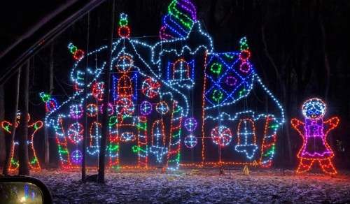Colorful holiday lights outline a whimsical gingerbread house surrounded by trees, creating a festive nighttime scene.