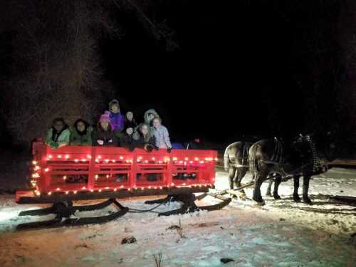 A group of children sits on a red, light-decorated sleigh pulled by two horses in a snowy, nighttime setting.