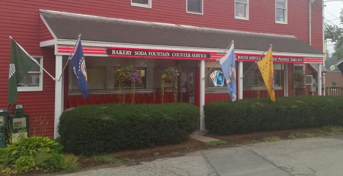 A red building with flags, featuring a bakery and soda fountain service, surrounded by greenery.