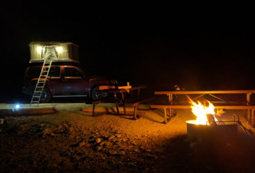 A truck with a rooftop tent parked near a campfire and picnic tables under a starry night sky.