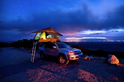 A car with a rooftop tent parked on a scenic overlook at dusk, with a colorful sky and distant lights.