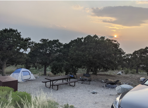 A campsite with a tent, picnic table, and car, surrounded by trees at sunset.