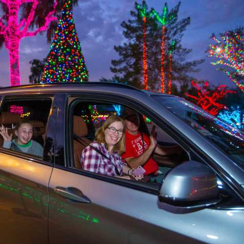 A family smiles and waves from a car, surrounded by colorful holiday lights in a festive outdoor setting.