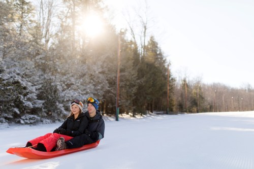 A man and woman sit together on a sled in a snowy landscape, surrounded by trees and sunlight.