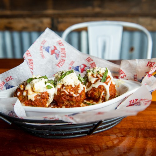 A plate of three meatballs topped with cheese and herbs, served in a basket lined with branded paper.