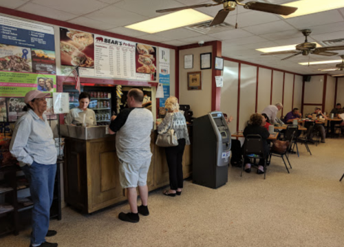 A busy restaurant interior with customers at the counter and seated at tables, featuring a menu board and an ATM.