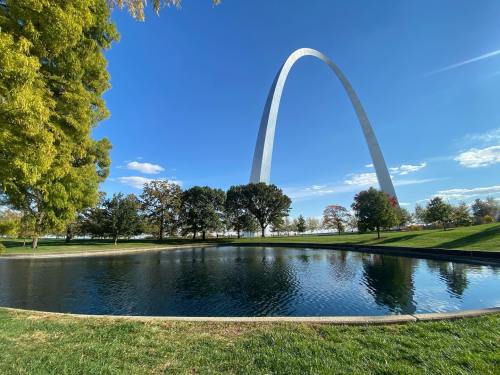 A tall arch monument rises above a serene pond, surrounded by trees and a clear blue sky.