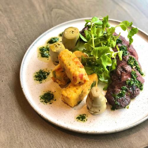 A plate featuring grilled meat, artichokes, polenta squares, and fresh greens drizzled with green sauce.