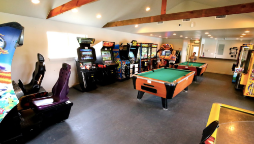 A game room featuring arcade machines, a pool table, and a racing game setup, with bright lighting and wooden beams.