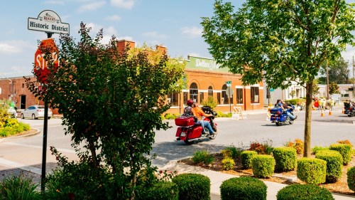 A scenic view of a historic district with greenery, a stop sign, and motorcycles riding through a charming town.