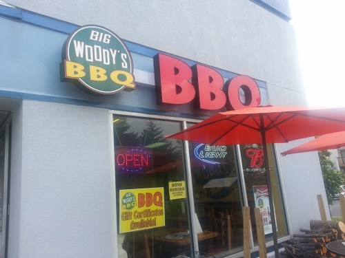 Sign for Big Woody's BBQ with "OPEN" and "Now Hiring" notices, featuring outdoor seating under red umbrellas.