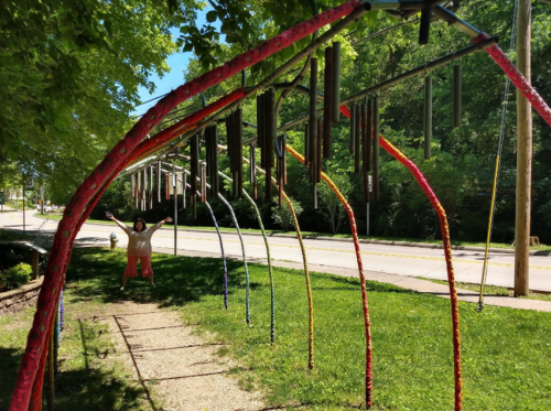A colorful archway of wind chimes with a person joyfully standing underneath on a sunny day.