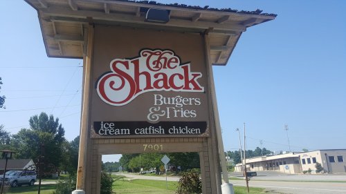 Sign for "The Shack" featuring burgers, fries, ice cream, catfish, and chicken, located on a sunny roadside.