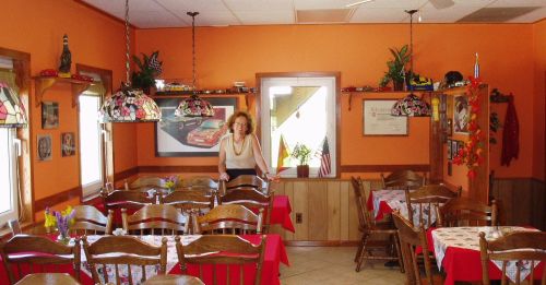 A woman stands in a cozy restaurant with orange walls, wooden tables, and colorful lamps, ready for diners.