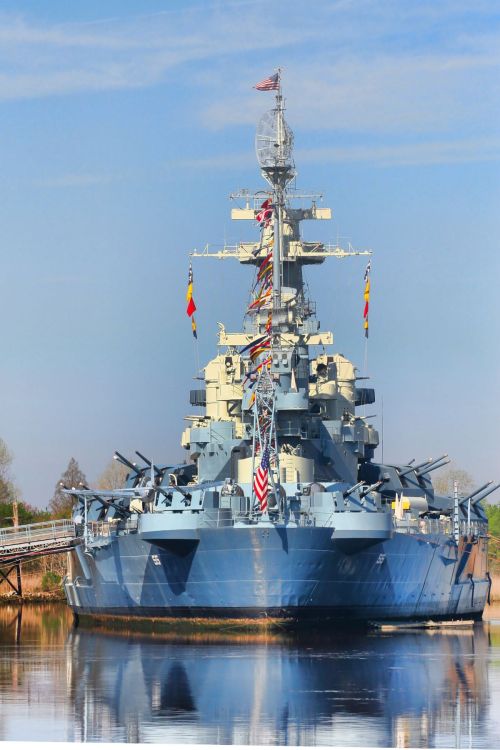 A large battleship docked on calm waters, adorned with flags and surrounded by trees under a clear blue sky.