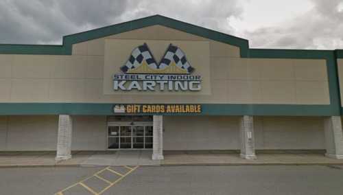 Exterior of Steel City Indoor Karting building with a large sign and "Gift Cards Available" banner. Cloudy sky above.