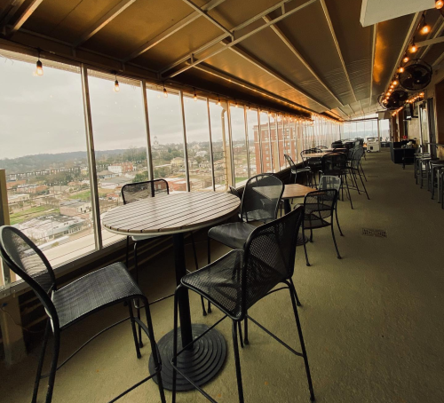 A rooftop patio with empty tables and chairs, offering a view of the city skyline through large windows.