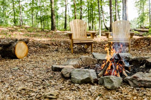 A cozy campfire surrounded by stones, with two wooden chairs nearby in a forested area.