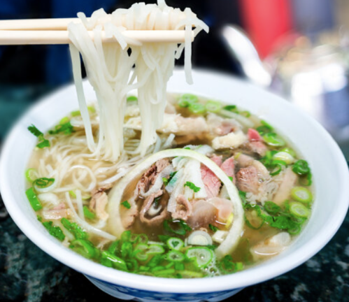 A bowl of steaming noodle soup with herbs, meat, and green onions, with chopsticks lifting noodles from the bowl.