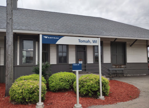 Sign for Tomah, WI Amtrak station in front of a building with a gray roof and landscaped bushes.