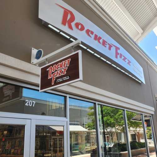 Sign for Rocket Fizz, a candy and soda shop, displayed on a storefront with large windows and a clear blue sky.