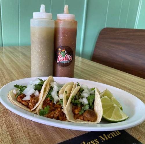 Three tacos topped with onions and cilantro, served with lime wedges and two bottles of sauce in the background.