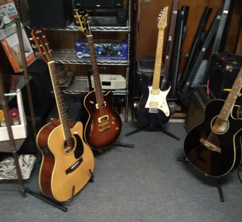 A collection of four guitars displayed on stands in a music room, with amplifiers and equipment in the background.