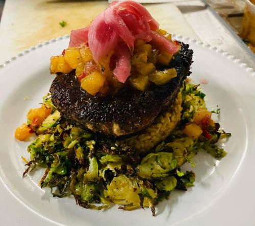 A beautifully plated dish featuring a grilled protein topped with pineapple salsa and pickled onions, served on a bed of sautéed Brussels sprouts.