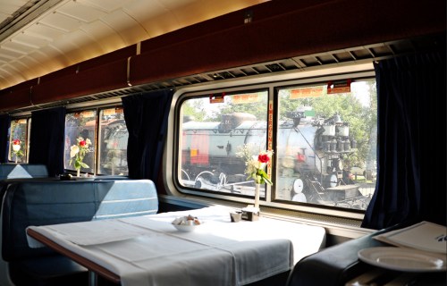 Interior of a train dining car with tables set, featuring large windows showing a steam locomotive outside.