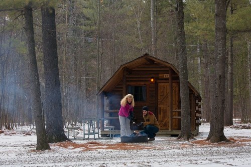 A couple prepares food over a fire pit outside a cozy cabin in a snowy forest. Pine trees surround the scene.