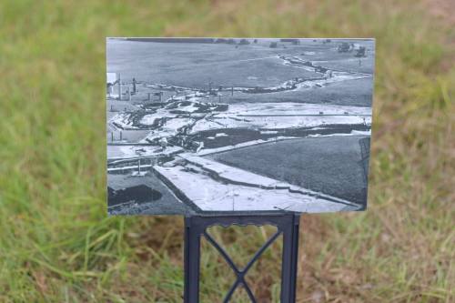 Black and white photo of a winding river through fields, displayed on a stand in a grassy area.