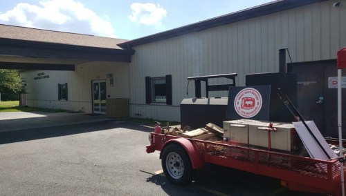 A red trailer with a smoker parked outside a building, with a sign on the trailer. Clear blue sky in the background.