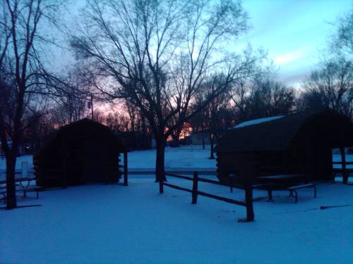 Two wooden cabins surrounded by snow, with trees and a colorful sunset in the background.