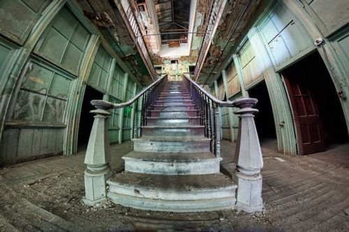 Abandoned building interior featuring a grand staircase, peeling walls, and dim lighting. Dust and debris are visible.