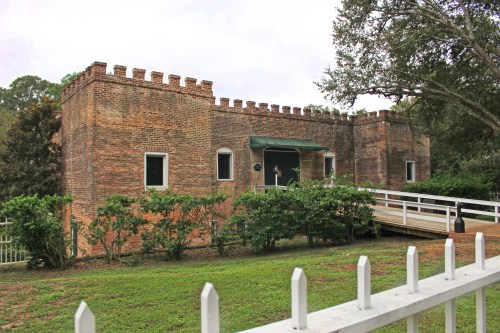 Historic brick building with a castle-like design, surrounded by greenery and a white picket fence.