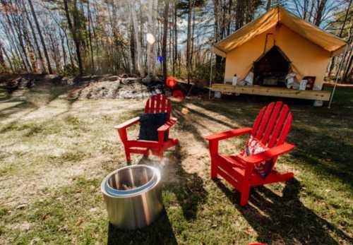 Two red Adirondack chairs sit by a fire pit in front of a glamping tent, surrounded by trees and sunlight.