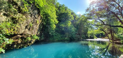 A serene blue river surrounded by lush green trees and rocky cliffs under a clear blue sky.