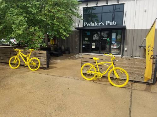 Two bright yellow bicycles are parked outside Pedaler's Pub, with a tree and building in the background.