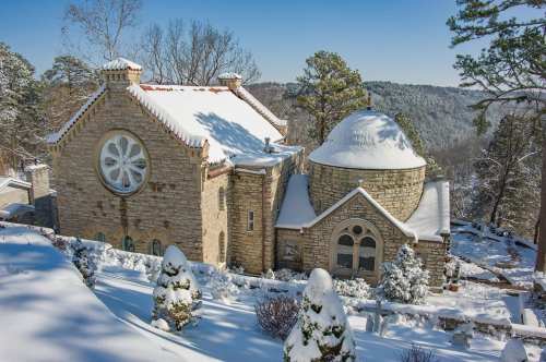 A snow-covered stone building with a round dome and large window, surrounded by trees and a winter landscape.