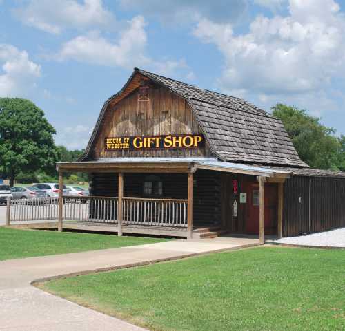 A rustic wooden gift shop with a sloped roof, surrounded by green grass and trees under a blue sky.