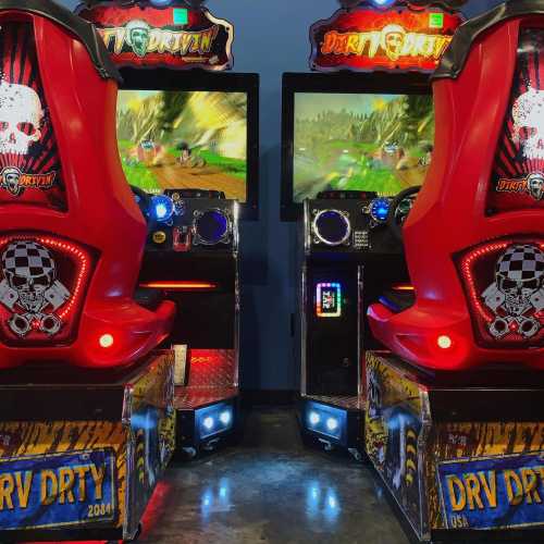 Two arcade racing game machines with vibrant screens and red racing seats, set in a dimly lit gaming area.