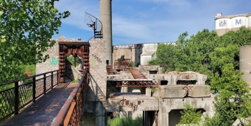 Abandoned industrial site with crumbling structures, overgrown vegetation, and a clear blue sky.