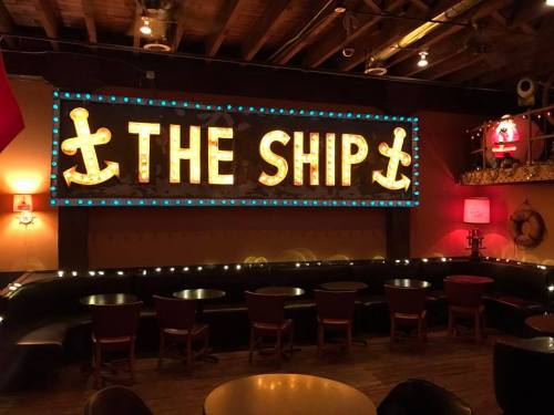 A cozy bar interior featuring a large illuminated sign that reads "THE SHIP" with anchor decorations.