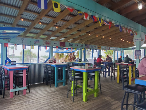 Colorful dining area with wooden tables and chairs, string lights, and nautical decorations, overlooking a waterfront view.