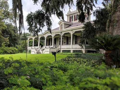 A historic mansion with a large porch, surrounded by lush greenery and trees, under a cloudy sky.