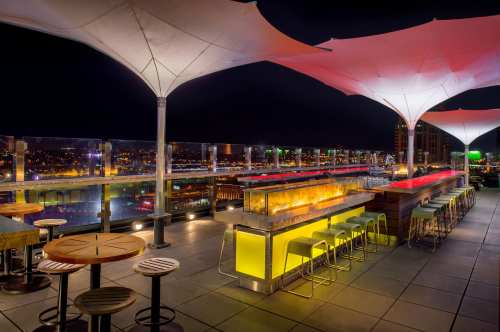 Rooftop bar at night with illuminated seating, fire features, and city skyline views under large white canopies.