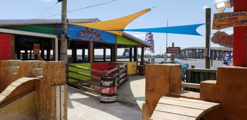 Colorful waterfront restaurant with a wooden deck, vibrant awnings, and a view of a bridge over the water.